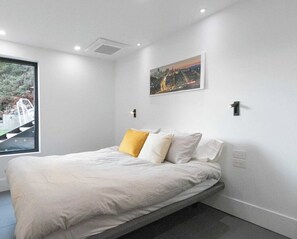 Ein modernes Schlafzimmer mit einem Bett, einem Stadtbildgemälde und einem Fenster mit Blick auf eine Straße und Grünflächen.