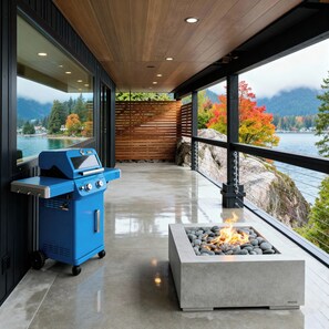 Eine moderne Außenküche mit eingebautem Grill und Feuerstelle mit Blick auf einen See und Herbstlaub.