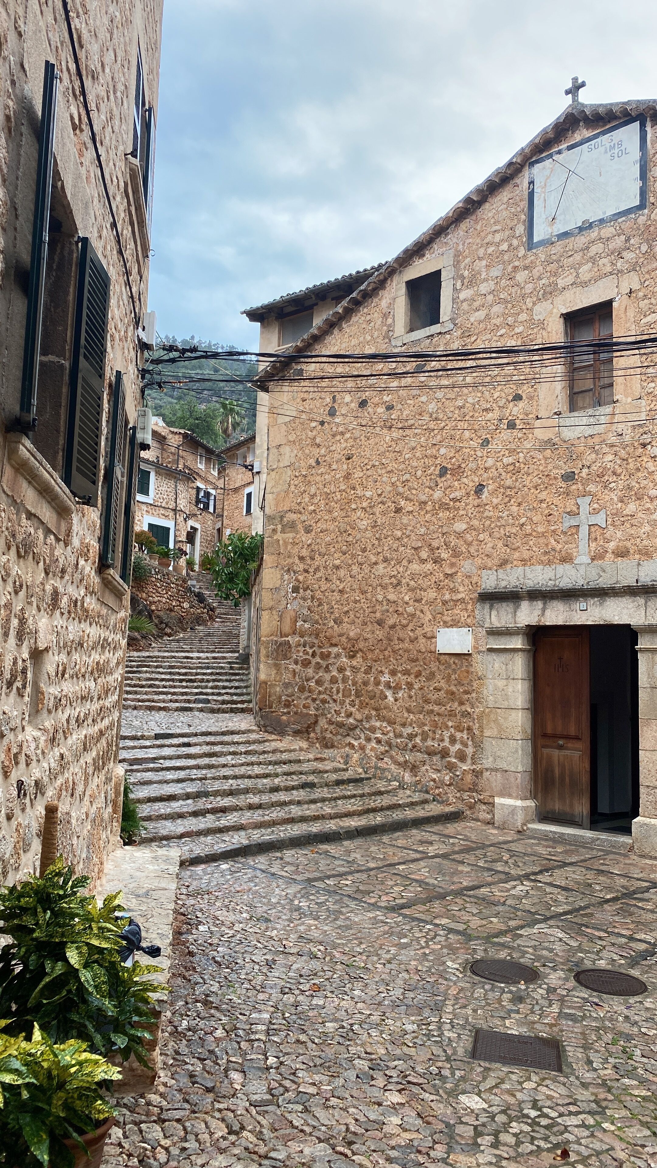 A cobblestone street leading to a stone building with a wooden door and a cross above it.
