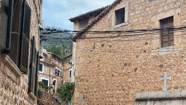 Eine Kopfsteinpflasterstraße führt zu einem Steinhaus mit einer Holztür und einem Kreuz darüber.