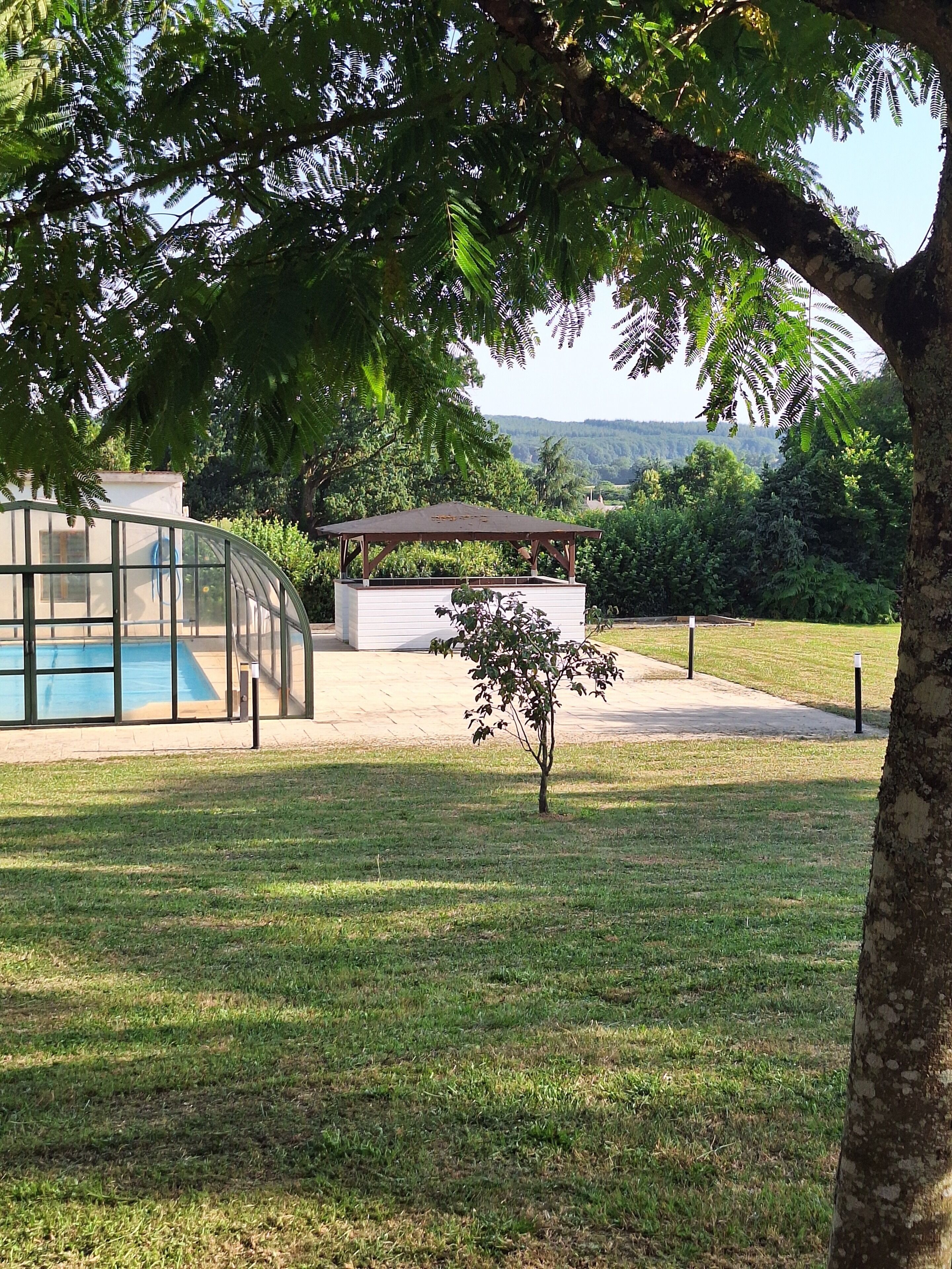 Un área de piscina con cerramiento de vidrio, con un quiosco, rodeada de césped y árboles.