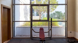 A room with a large window, a bench, and a weightlifting station. - Stylish Studio in DAMAC Hills with Pool, Gym & Balcony (Damac hills)