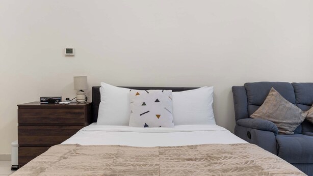 A bedroom with a bed, a bedside table, a lamp, a chair, and a wall-mounted thermostat. - Stylish Studio in DAMAC Hills with Pool, Gym & Balcony (Damac hills)