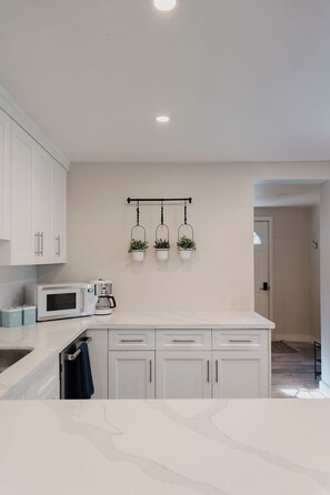 Une cuisine moderne avec des meubles blancs, un plan de travail blanc, un four à micro-ondes, une machine à café et trois pots de fleurs suspendus.