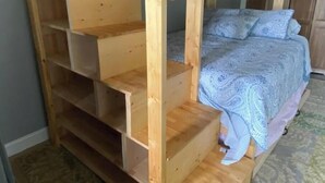Un lit superposé avec une échelle en bois et une étagère inférieure, une couvre-lit à motifs et un sol recouvert d’une moquette.