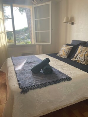 Ein ordentlich bezogenes Bett mit einem zusammengerollten Handtuch, ein Fenster mit Vorhängen und eine Wandlampe.