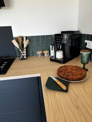 Et køkken med kaffemaskine, træredskaber, krukker, en kage og et grønt glas.
