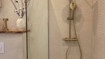 A modern bathroom with a glass shower enclosure, a gold showerhead, and a vase with cotton branches on the vanity.