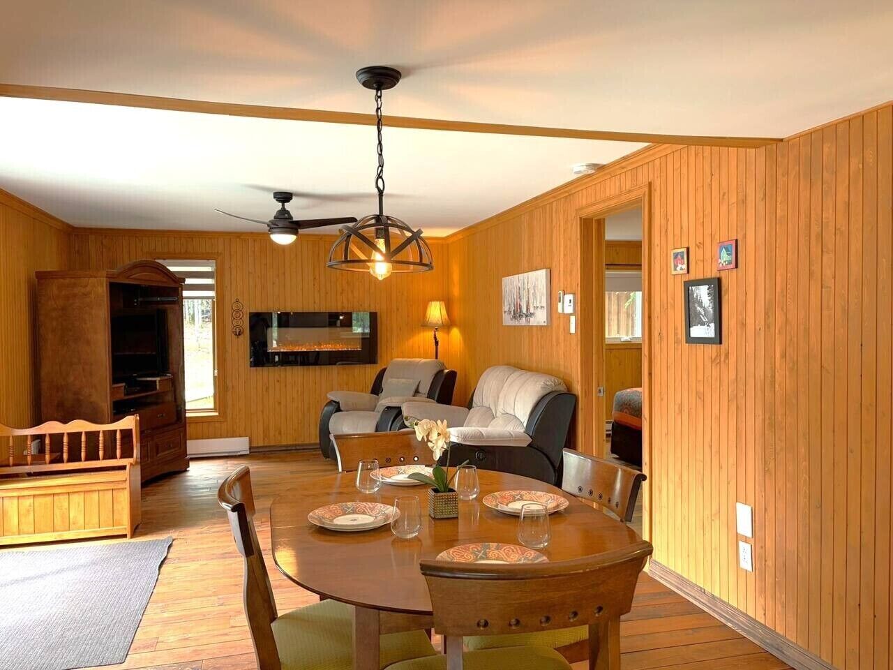 Um quarto com painéis de madeira, uma mesa de jantar posta para quatro pessoas, um sofá e uma televisão.