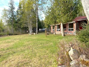 A rustic cabin with a red roof, surrounded by a grassy yard and dense forest.