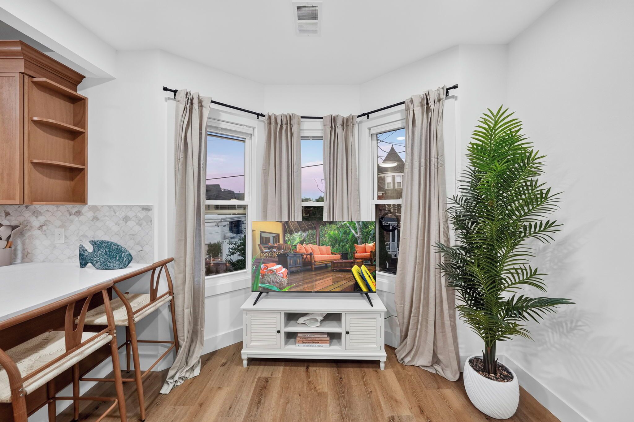 A corner room with a TV, wooden furniture, a potted plant, and a view of a house through the windows.