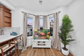 Habitación de esquina con televisor, muebles de madera, una planta en maceta y vista a una casa a través de las ventanas.