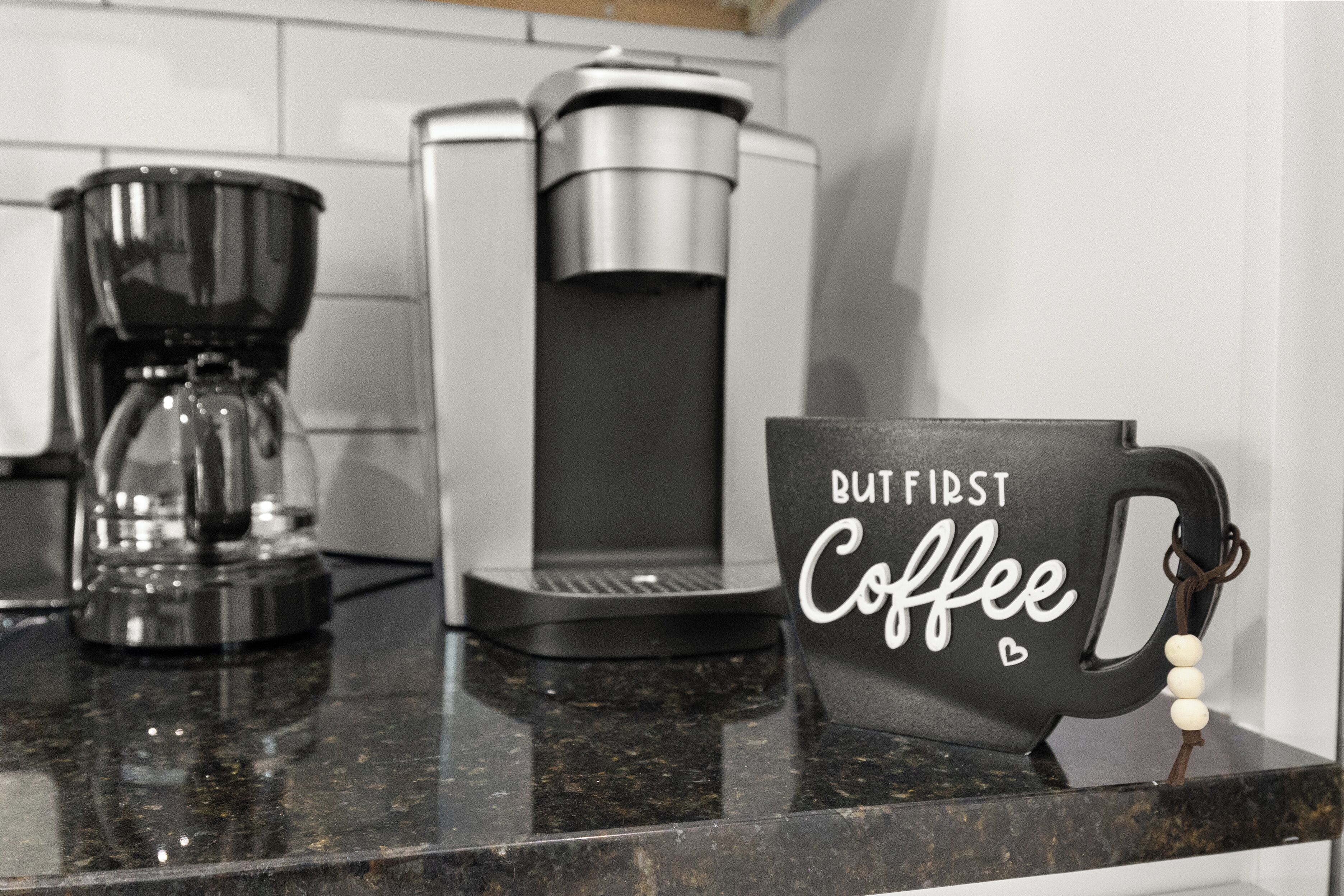 Un piano di lavoro in cucina con una macchina da caffè, un macinino e una tazza con la scritta 'BUT FIRST Coffee'.