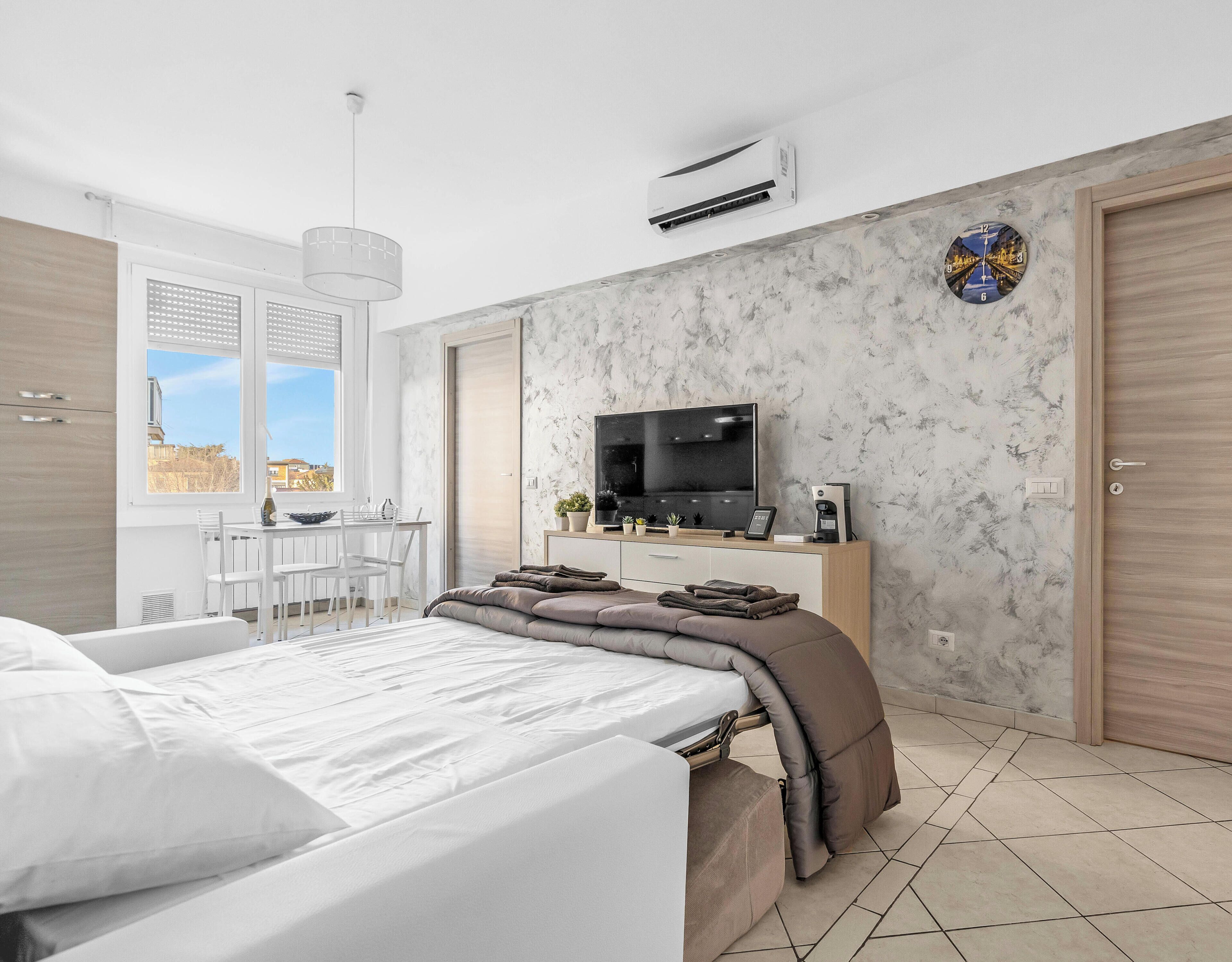 A modern bedroom with a bed, a TV, a desk, and a view of the city.