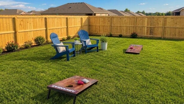 A backyard with a wooden fence, a lawn, two blue chairs, a small table, and a wooden board game.