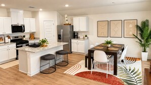 A modern kitchen with white cabinetry, a central island with stools, a dining area with a dark wooden table and white chairs, and a large potted plant.