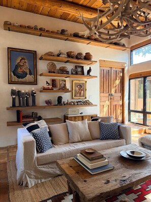 A cozy living room with a wooden ceiling, shelves with pottery, a sofa with patterned cushions, and a rustic coffee table. - Hacienda Coco: An Artful, Dog-Friendly Retreat Near the Opera, Plaza and Tesuque (Santa Fe)
