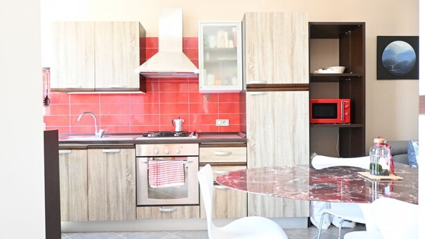 A modern kitchen with wooden cabinets, a red tiled backsplash, a gas stove, a microwave, and a dining area with a glass table and white chairs.