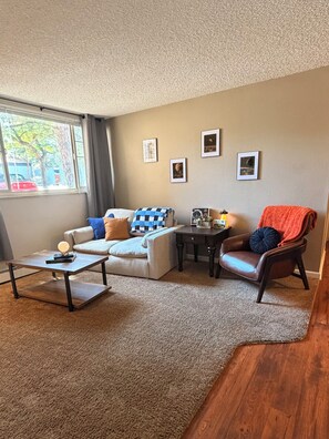 Ein Wohnzimmer mit einer Couch, einem Couchtisch, einem Sessel und einem kleinen Tisch mit einer Lampe. An den Wänden befinden sich Bilderrahmen und ein Fenster mit Blick ins Freie.