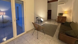 A spacious living area with a sliding glass door, a sofa, a chair, a lamp, and a wooden cabinet. - Village house nestled among ponds, scrubland, and a wild beach (La Palme)