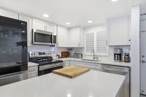 A modern kitchen with white cabinetry, stainless steel appliances, a central island with a cutting board, and a coffee maker. - Oceanfront Daytona Beach Shores Condo - King Suite - Sleeps 6 (Daytona Beach Shores)