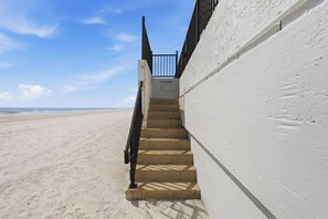 Eine Betonstufenanlage führt zu einem verschlossenen Eingang, neben einem sandigen Strand mit blauem Himmel.
