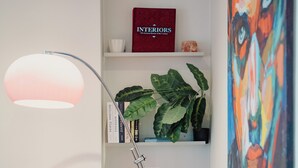 Ett modernt rum med en svart läderfåtölj, en golvlampa, en bokhylla med böcker och en växt, en röd låda märkt med texten ”INREDNING” och en färgstark, abstrakt målning.