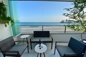 Un balcón con vista a la playa y al océano. Hay dos sillas y una mesita con una botella y un vaso.