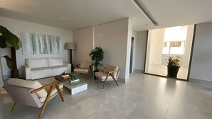 Un salon moderne avec un canapé blanc, deux fauteuils, une table basse et un lampadaire. Une grande fenêtre laisse entrer la lumière naturelle et offre une vue sur l’extérieur.