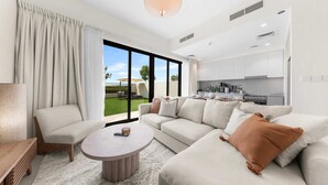 Una sala de estar moderna con sofá, sillón y mesa de centro, una zona de cocina y grandes puertas correderas de vidrio hacia un jardín.
