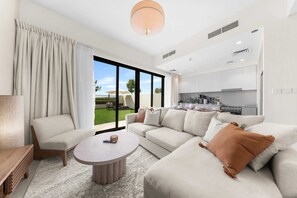 A modern living room with a sofa, armchair, and coffee table, a kitchen area, and large sliding glass doors to a garden. - Free Housekeeping Visits During Your Stay - Stayshort - Exceptional 3 Bedroom Villa in Emaar South That Sleeps 8 (Dubai)