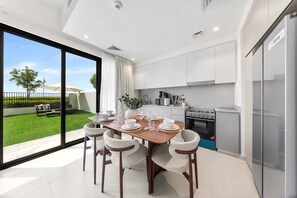 A modern kitchen with a dining area, large windows, and a view of a green lawn. - Free Housekeeping Visits During Your Stay - Stayshort - Exceptional 3 Bedroom Villa in Emaar South That Sleeps 8 (Dubai)