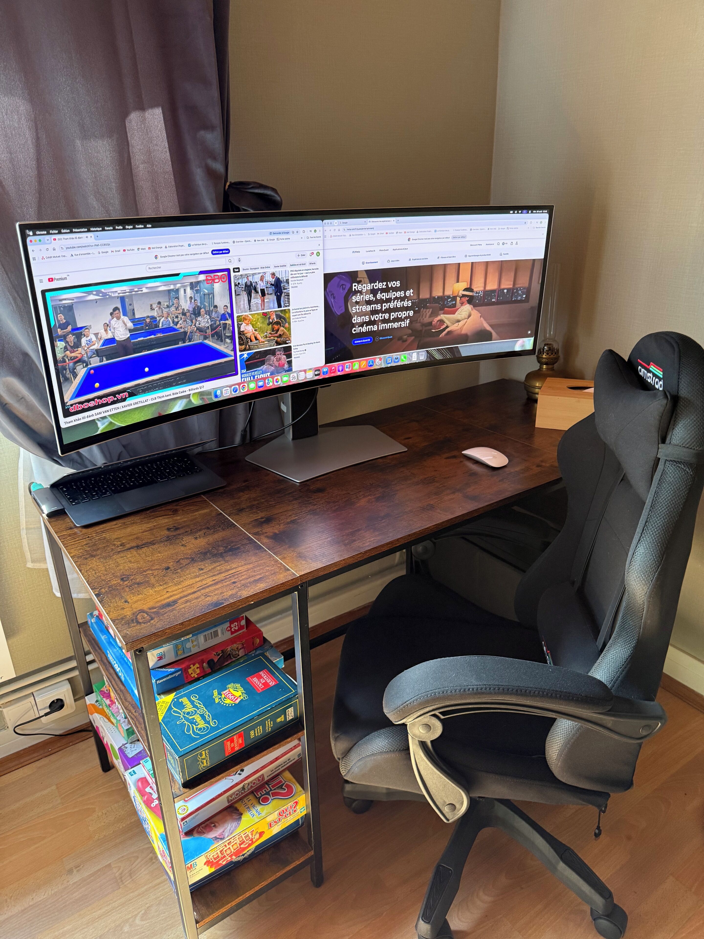 Dubbele monitoren op een houten bureau, met een gaming stoel, een laptop en een boekenplank vol gezelschapsspellen.