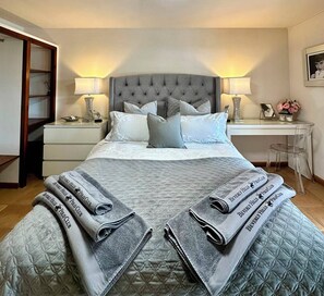 Un lit bien fait, avec une couverture grise et des oreillers, une table de chevet avec une lampe, un bureau avec une chaise et un tableau encadré au mur.