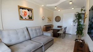 A living room with a grey sofa, a dining table with chairs, a small kitchen area, and a potted plant. - SHB Paraíso na Praia de Pitangueiras (Guarujá)