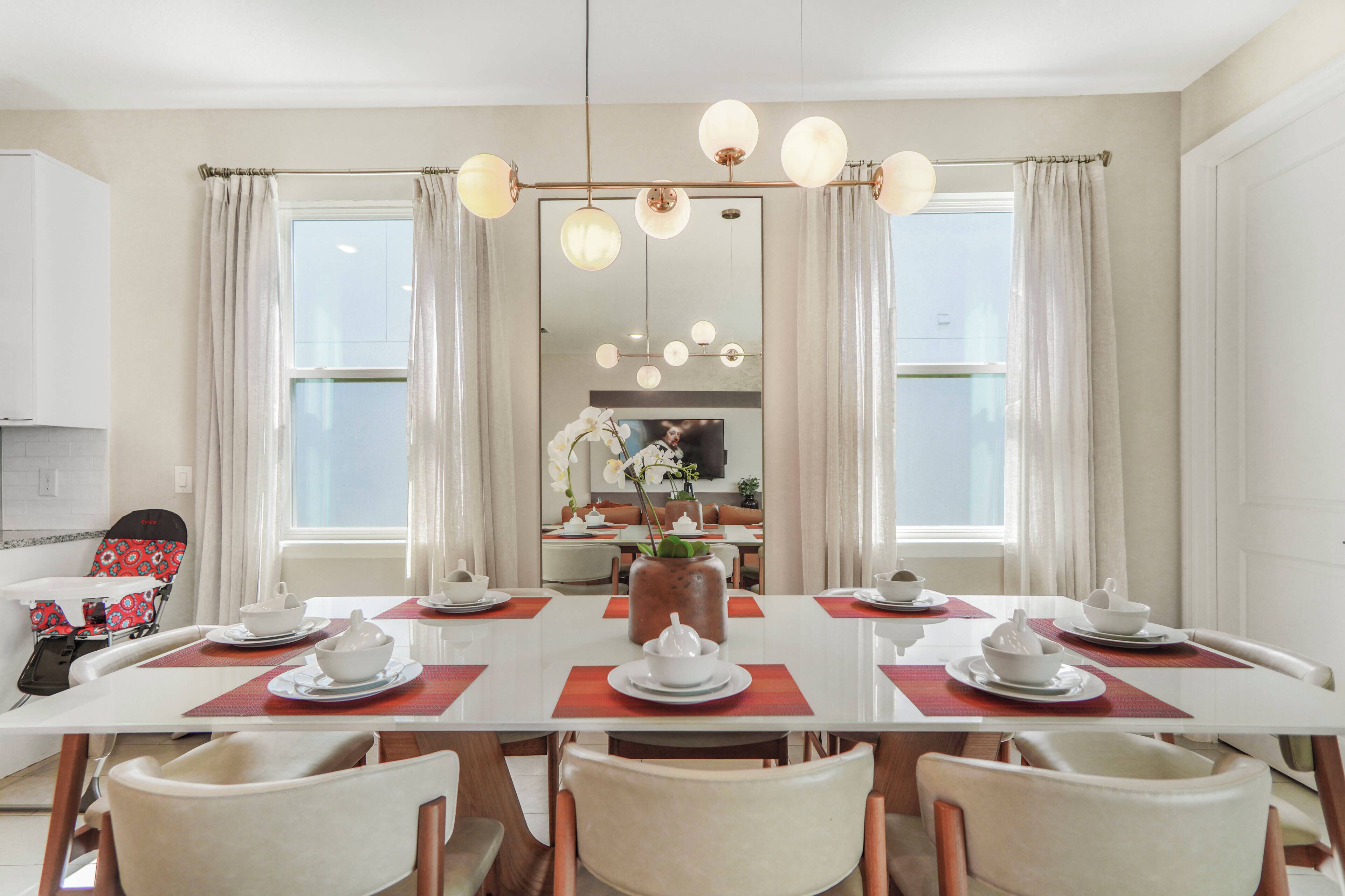 A dining area with a white table set for six, surrounded by chairs, with a kitchen visible in the background.