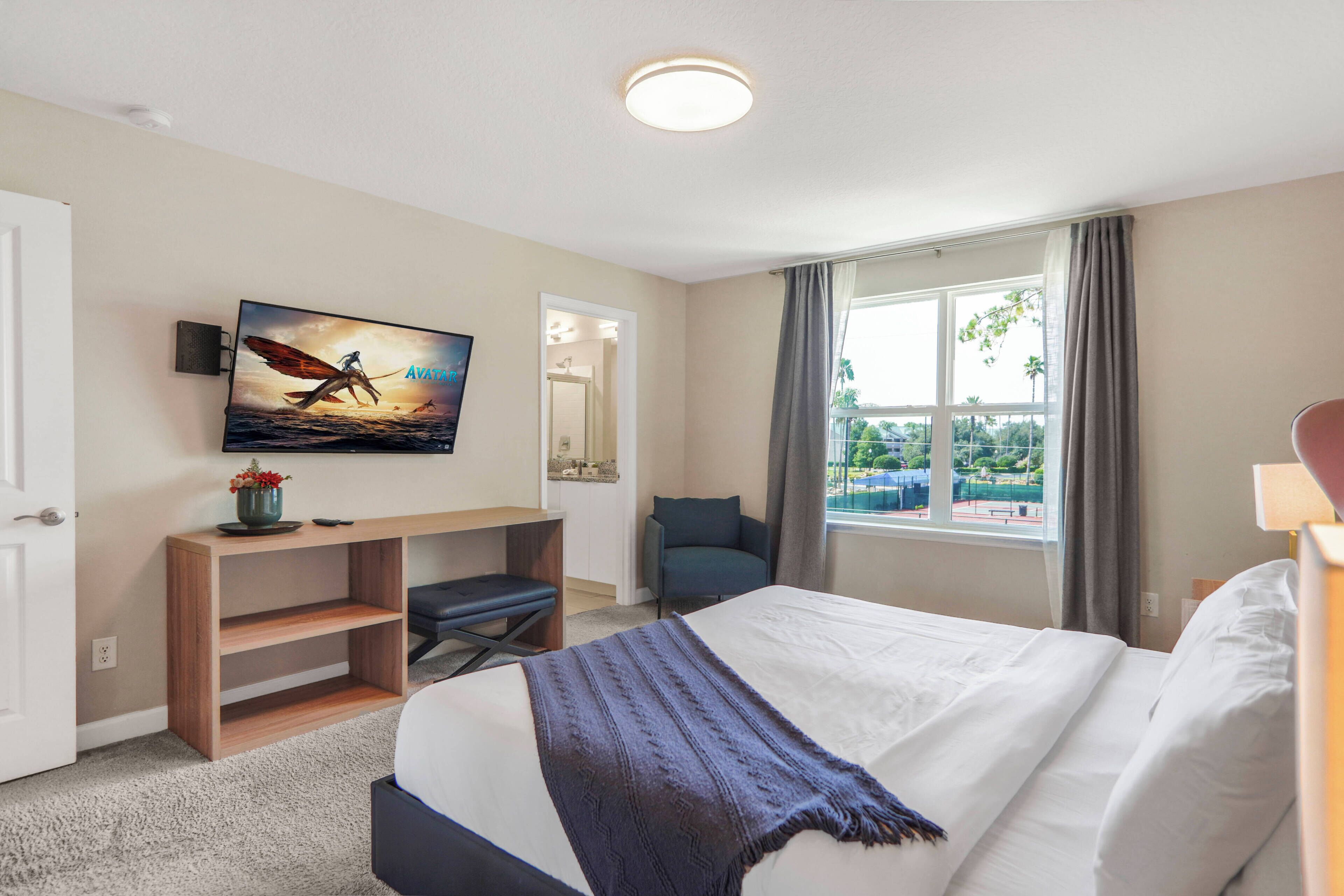 A hotel room with a bed, a desk, a chair, a TV mounted on the wall, and a window with a view.