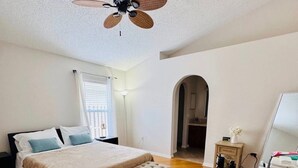 A bedroom with a bed, a bench, a ceiling fan, a window with blinds, and a door leading to another room.