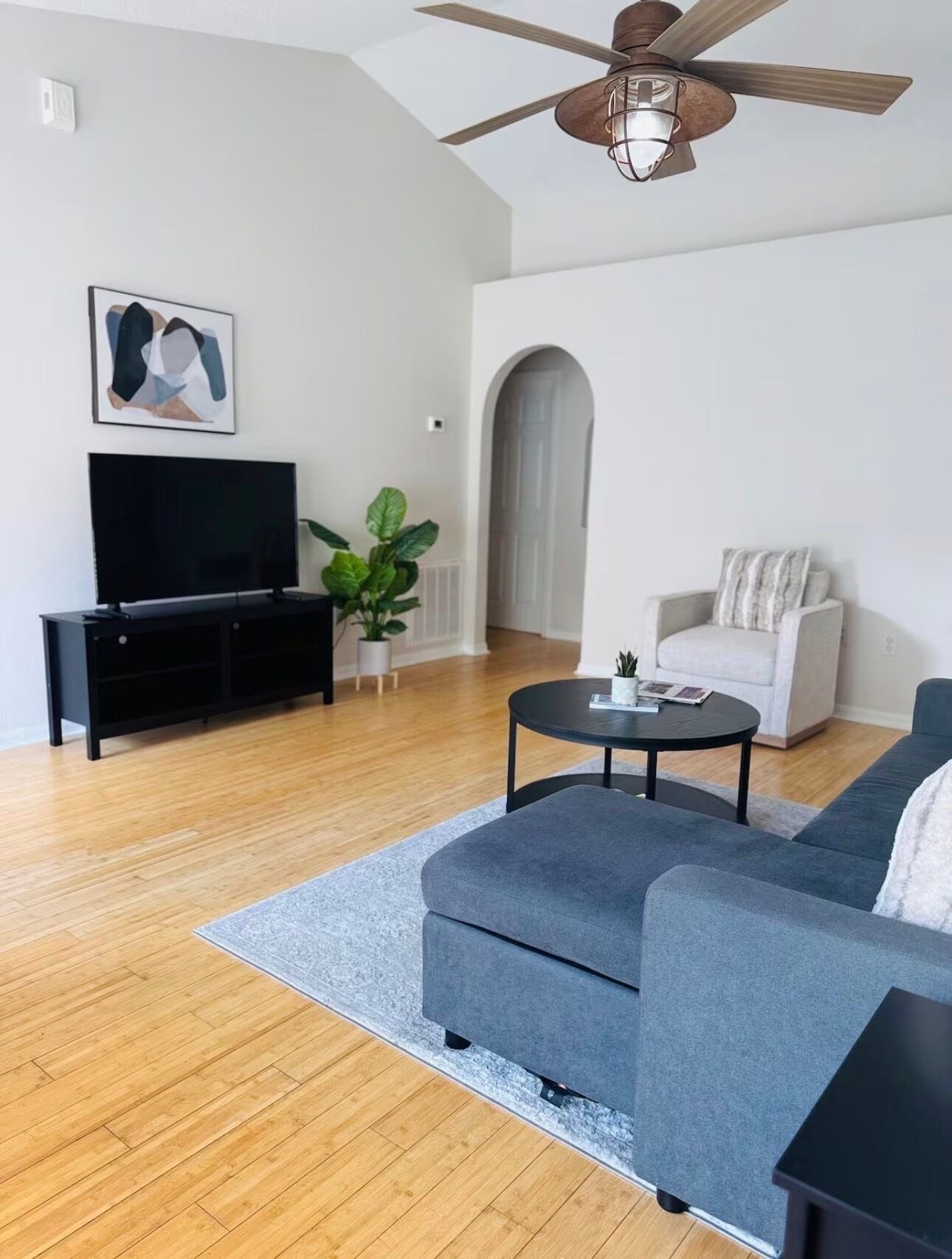 A modern living room with a flat-screen TV, a grey sofa, a coffee table, and a ceiling fan.