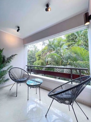 A balcony with two chairs and a small table, a large window with a view of greenery, and a ceiling light fixture. - 4-bedroom Poblado Provenza walk to lleras (Medellín)