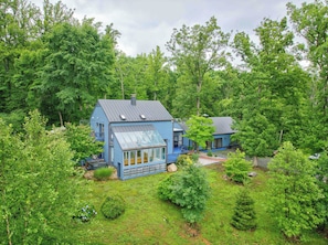 A blue house with a large glass window and a metal roof, surrounded by dense green trees. - Luxury Blue Ridge Retreat - Hot Tub - 15 Min to Early Mountain Vineyard (Stanardsville)