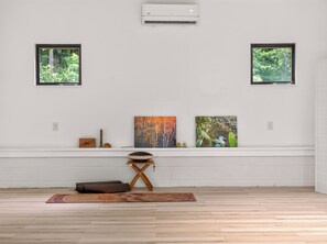 A room with a white wall, two windows with views of greenery, a wooden floor, a small table with a cushion, and a wall-mounted air conditioner. - Luxury Blue Ridge Retreat - Hot Tub - 15 Min to Early Mountain Vineyard (Stanardsville)