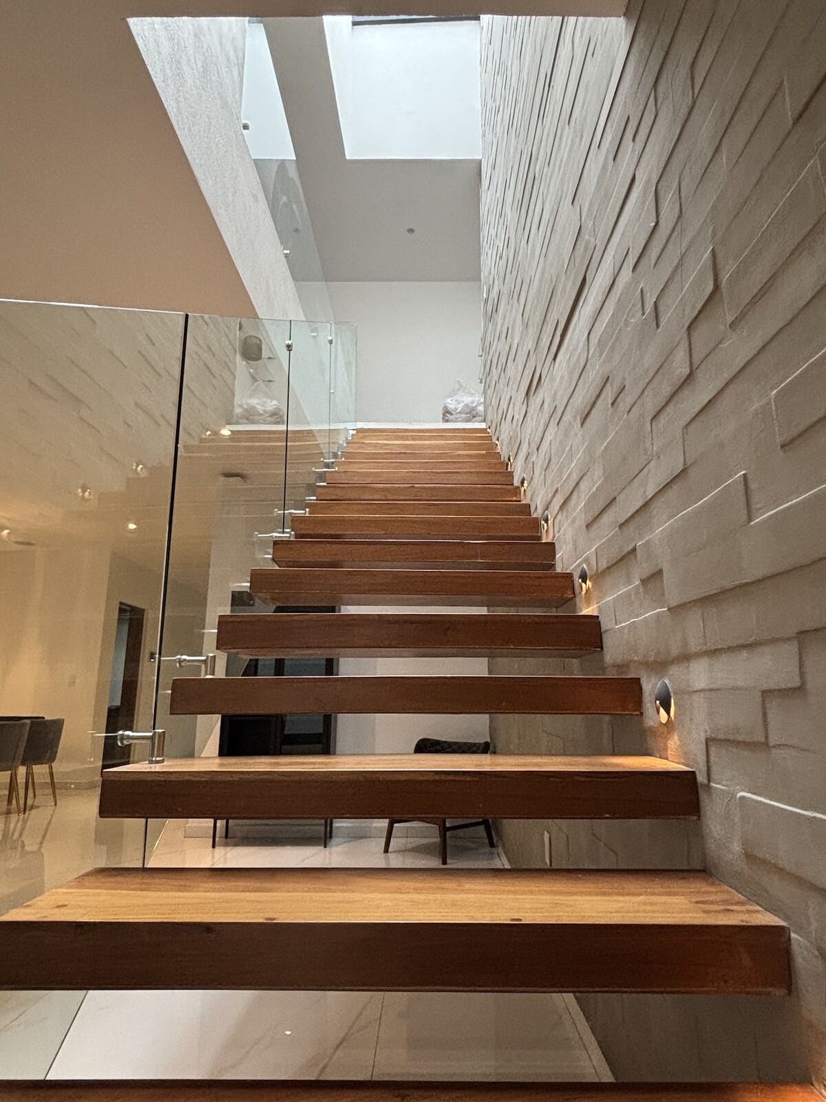 Una escalera moderna con escalones de madera y barandillas de cristal, que conduce a un techo alto con tragaluces.