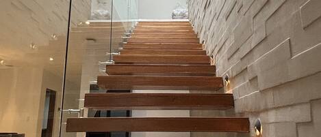 Una escalera moderna con escalones de madera y barandillas de cristal, que conduce a un techo alto con tragaluces.