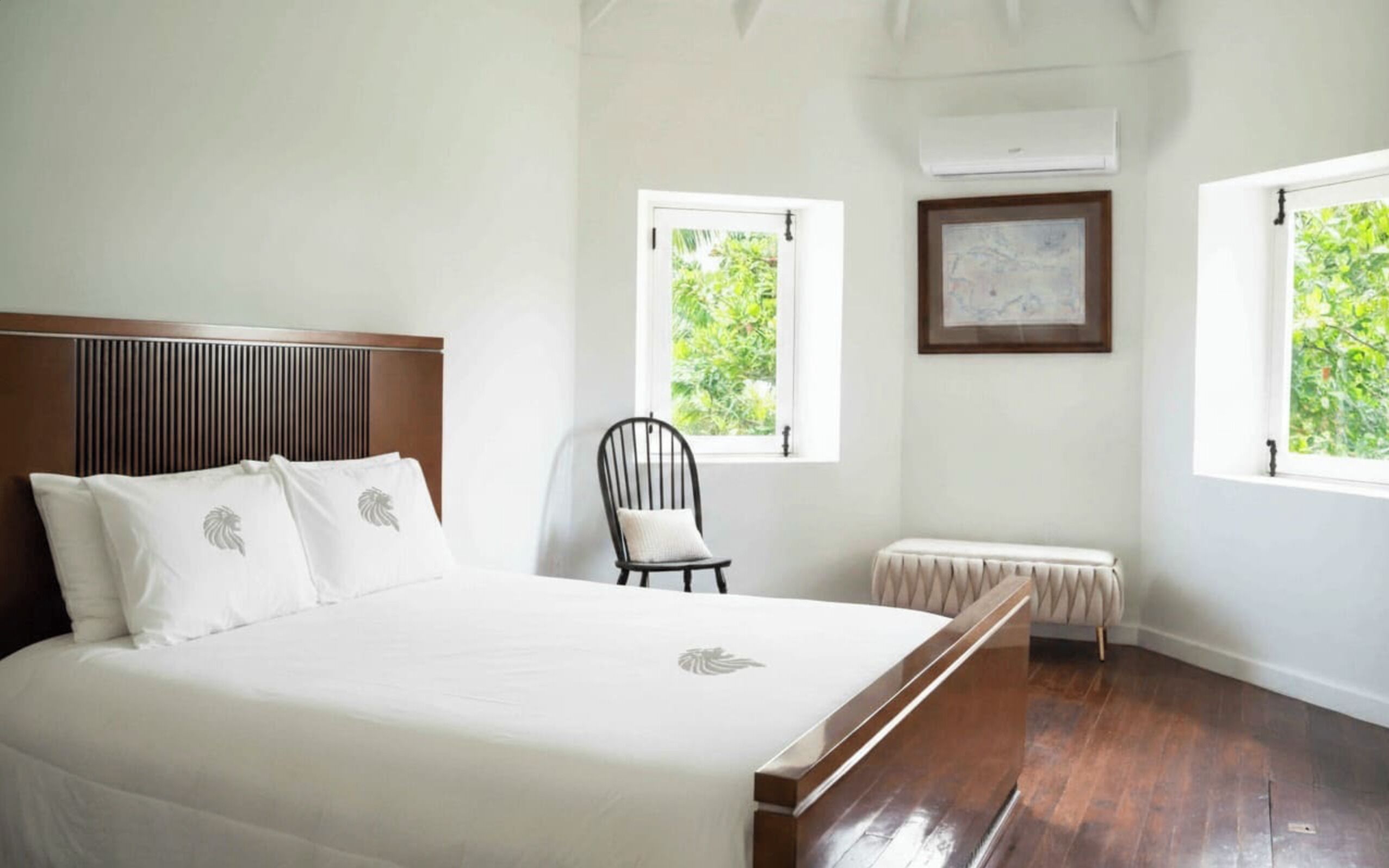 Quarto com uma cama grande, uma cadeira, um banco e duas janelas com vista para o verde.
