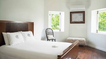 Quarto com uma cama grande, uma cadeira, um banco e duas janelas com vista para o verde.