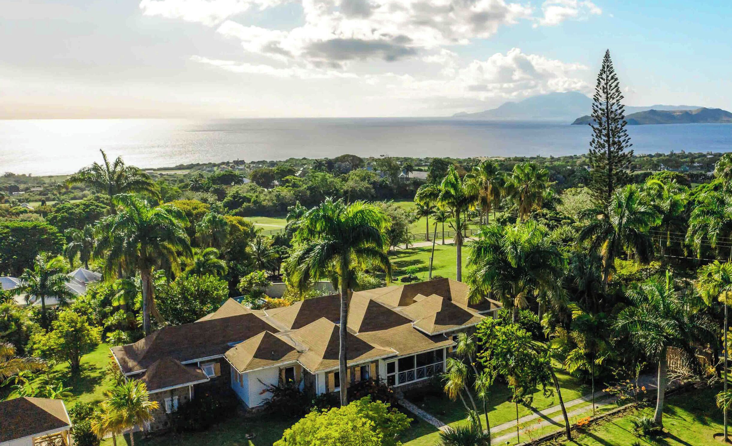 A large house with a sloped roof, surrounded by lush greenery and palm trees, overlooking a body of water and distant mountains.