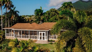 Um edifício de dois andares com varanda, rodeado por vegetação tropical e montanhas ao fundo.