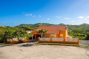 A two-story house with a red-tiled roof, surrounded by a white picket fence, set against a backdrop of green hills and a clear blue sky.