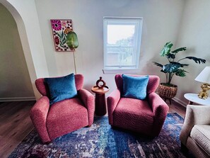 Une pièce comprenant deux fauteuils, une petite table d’appoint, une plante en pot, une fenêtre avec des stores et une œuvre d’art colorée accrochée au mur.